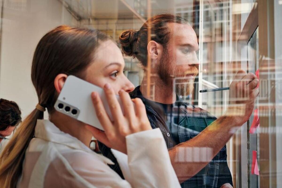 Młoda kobieta rozmawia przez telefon w biurze. W tle mężczyzna robi notatki na kartkach
