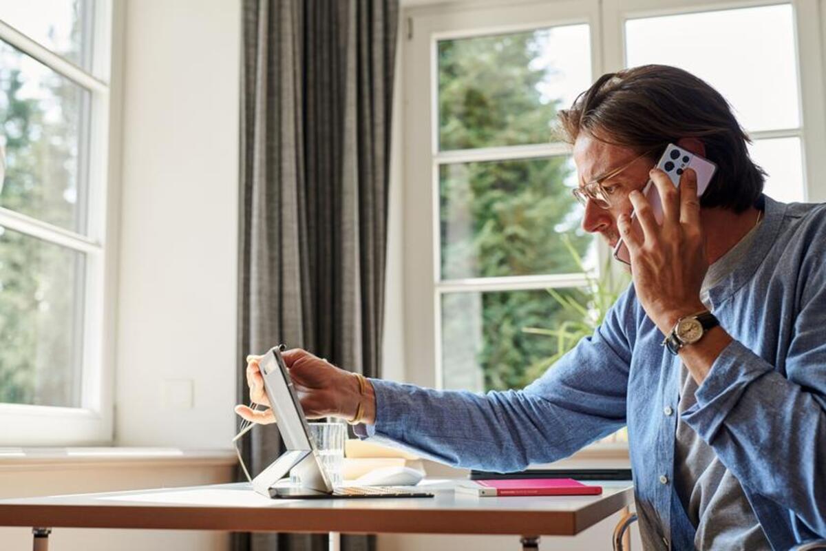 Mężczyzna w domowym biurze rozmawia przez telefon i jednocześnie korzysta z tabletu