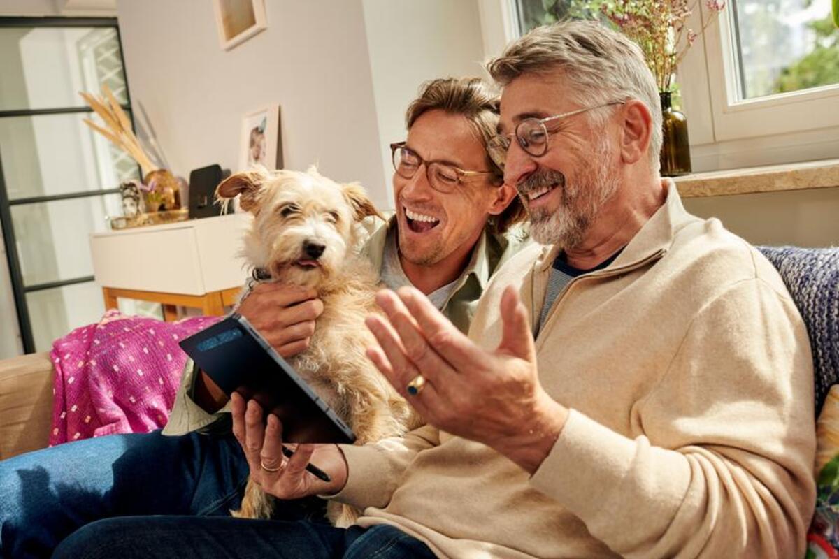 Dwóch uśmiechniętych mężczyzn siedzi na kanapie. Jeden trzyma psa, a drugi tablet.